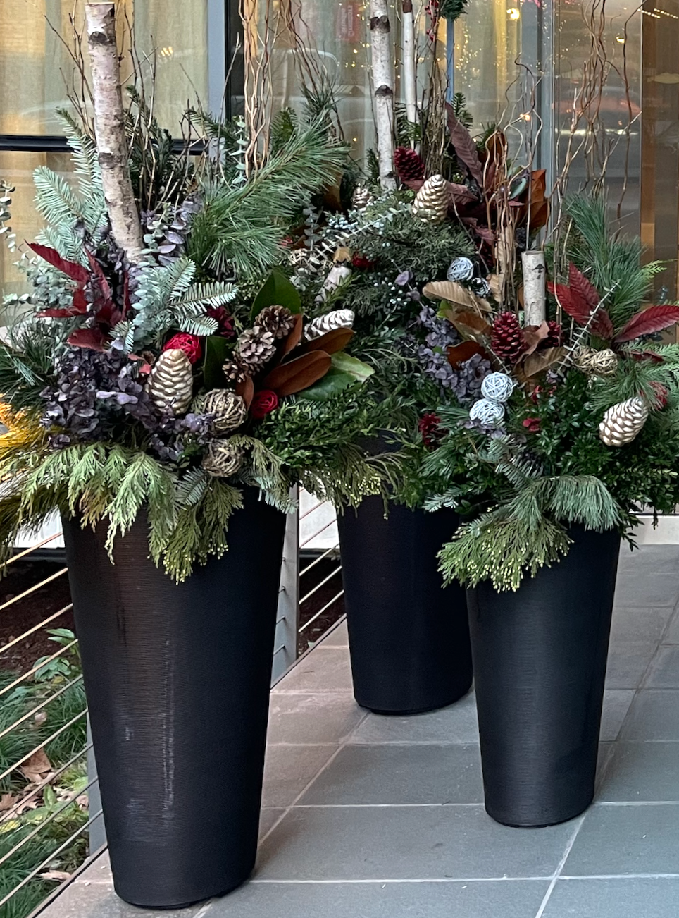 Two black pots with holiday decorations. One pot contains a wreath of fresh cut seasonal greens, ornament balls, and pinecones.