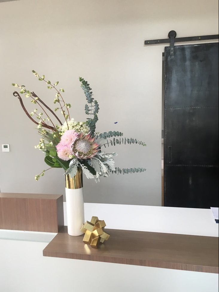 A floral arrangement with a mix of flowers and greenery in a white vase, placed on a desk with a gold-colored gift tag, set against a neutral wall in what appears to be a corporate or office setting.